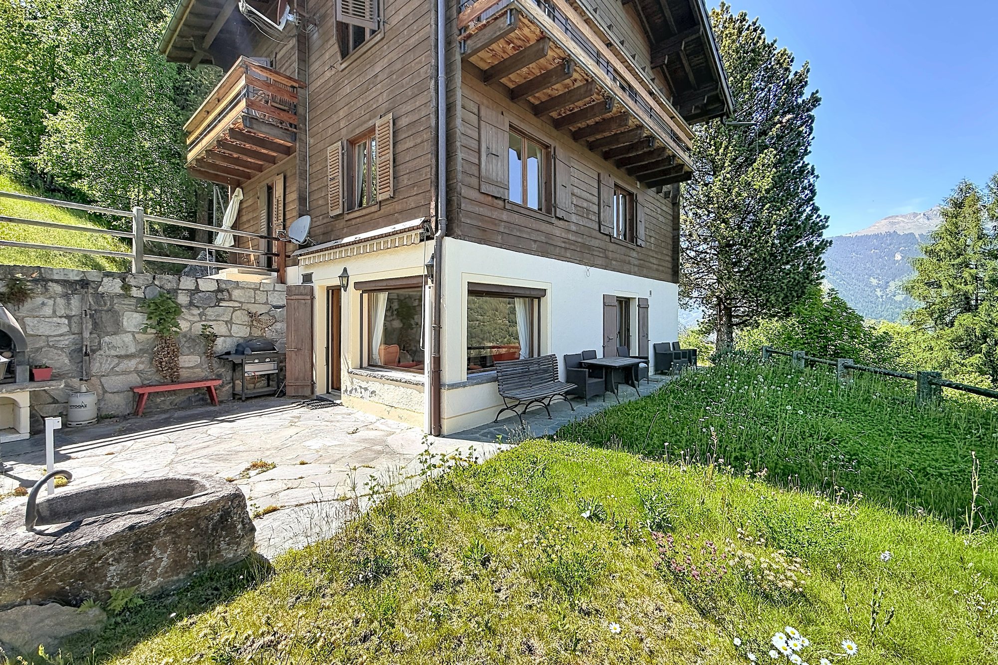 Le chalet vu du jardin : la terrasse en pierres naturelles, le barbecue et la vue sur la vallée.